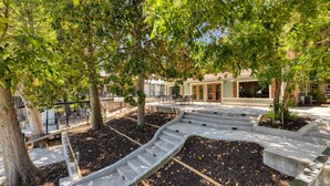 A landscaped outdoor area with a curved concrete pathway, trees, and a building with large windows in the background.