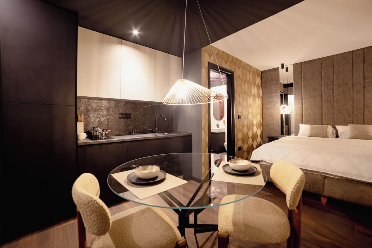 A modern hotel room with a glass dining table, two chairs, a bed, a small kitchenette, and a unique textured wall design.
