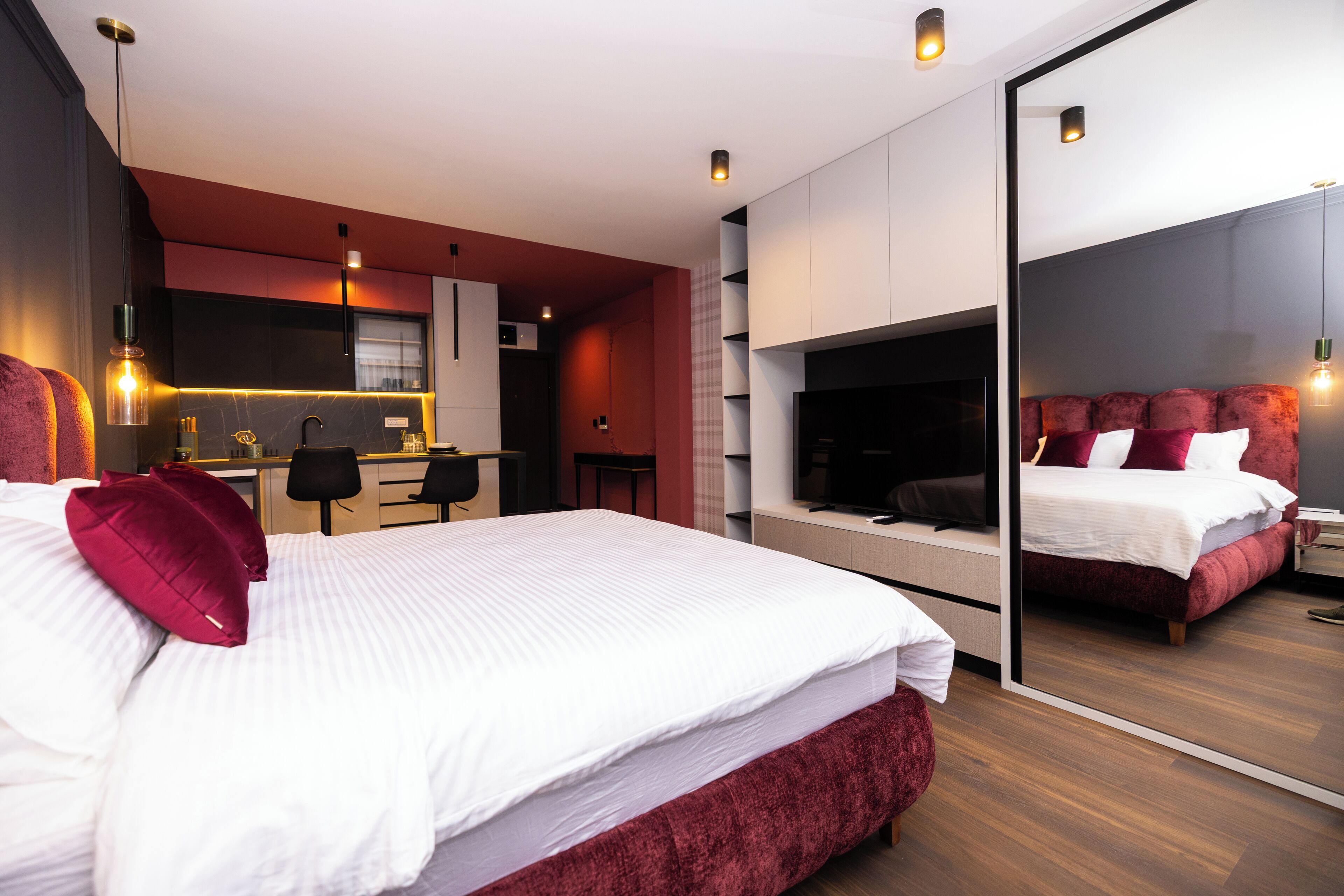 A modern hotel room with a large bed, a TV, a desk, and a kitchen area.