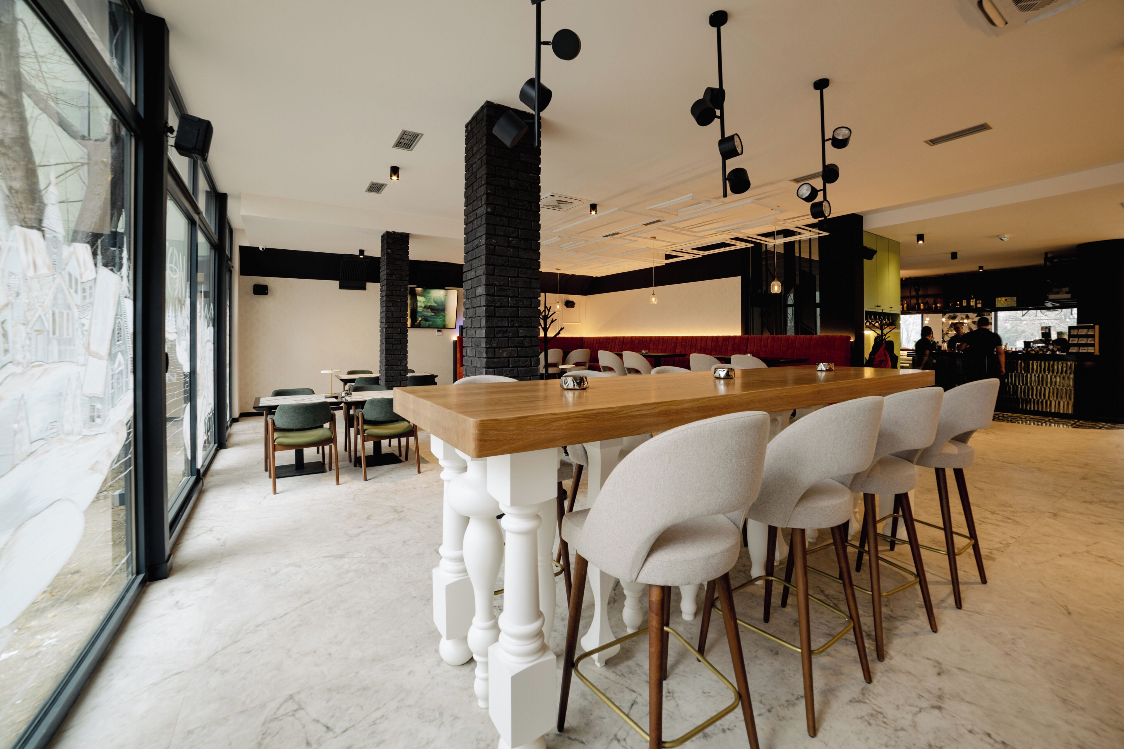 Un restaurante moderno con una larga mesa de madera y sillas blancas, ventanas amplias y un pilar de ladrillo negro.