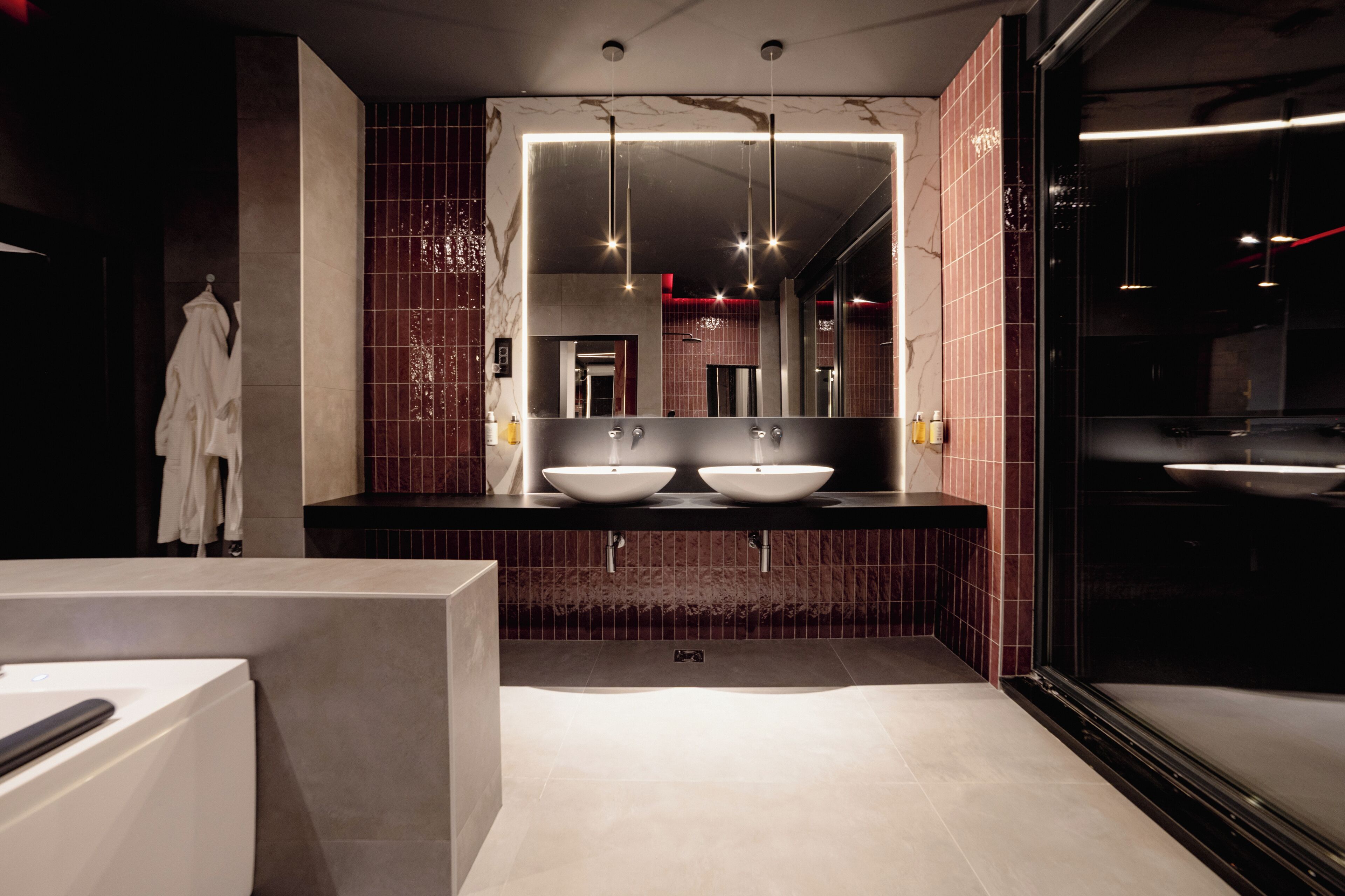 Une salle de bain moderne avec deux vasques, un grand miroir et un mur recouvert de carreaux foncés.