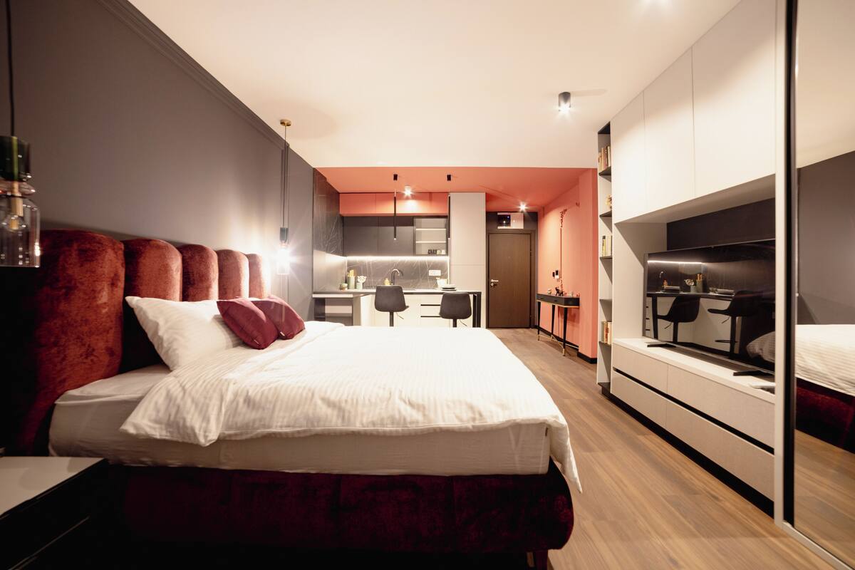 A modern bedroom with a large bed, a desk, and a kitchenette in the background.