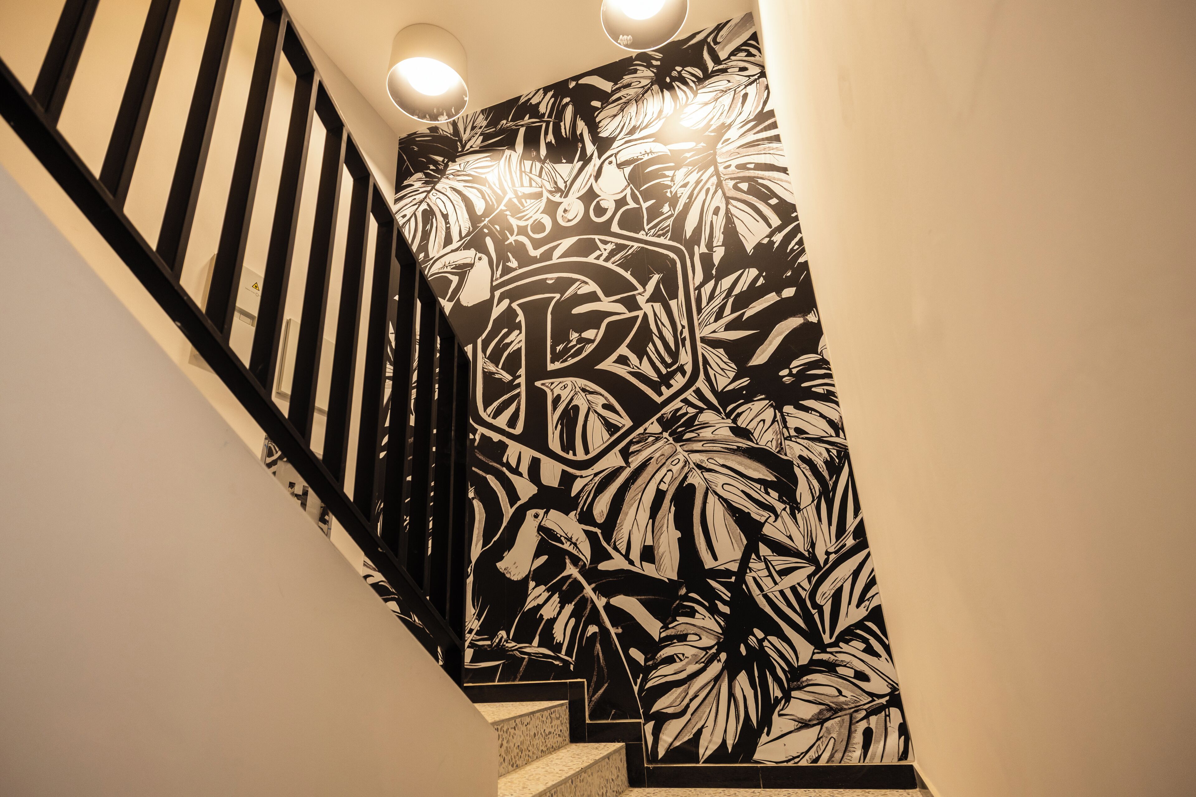 A staircase with a black and white tropical leaf pattern on the wall, a black railing, and three wall-mounted lights.