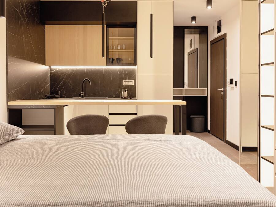 A modern hotel room with a bed, a desk with a chair, a small kitchenette, and a wardrobe.