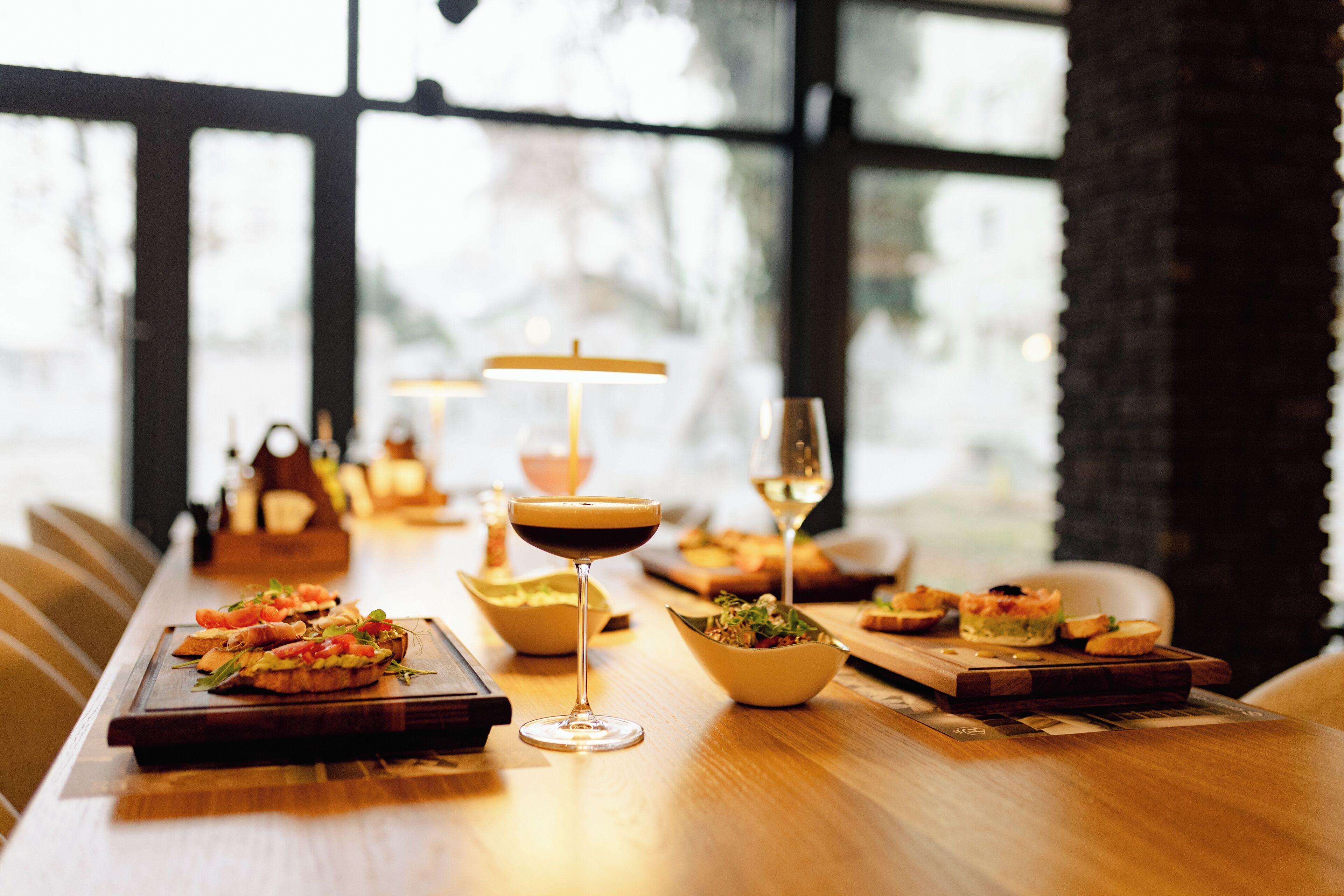 A long wooden table with various dishes and drinks, set in a well-lit room with large windows.