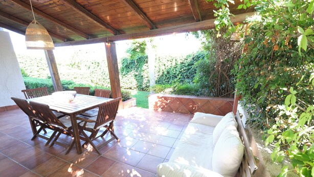 Um pátio externo com mesa e cadeiras de madeira, um sofá branco e um candeeiro suspenso.