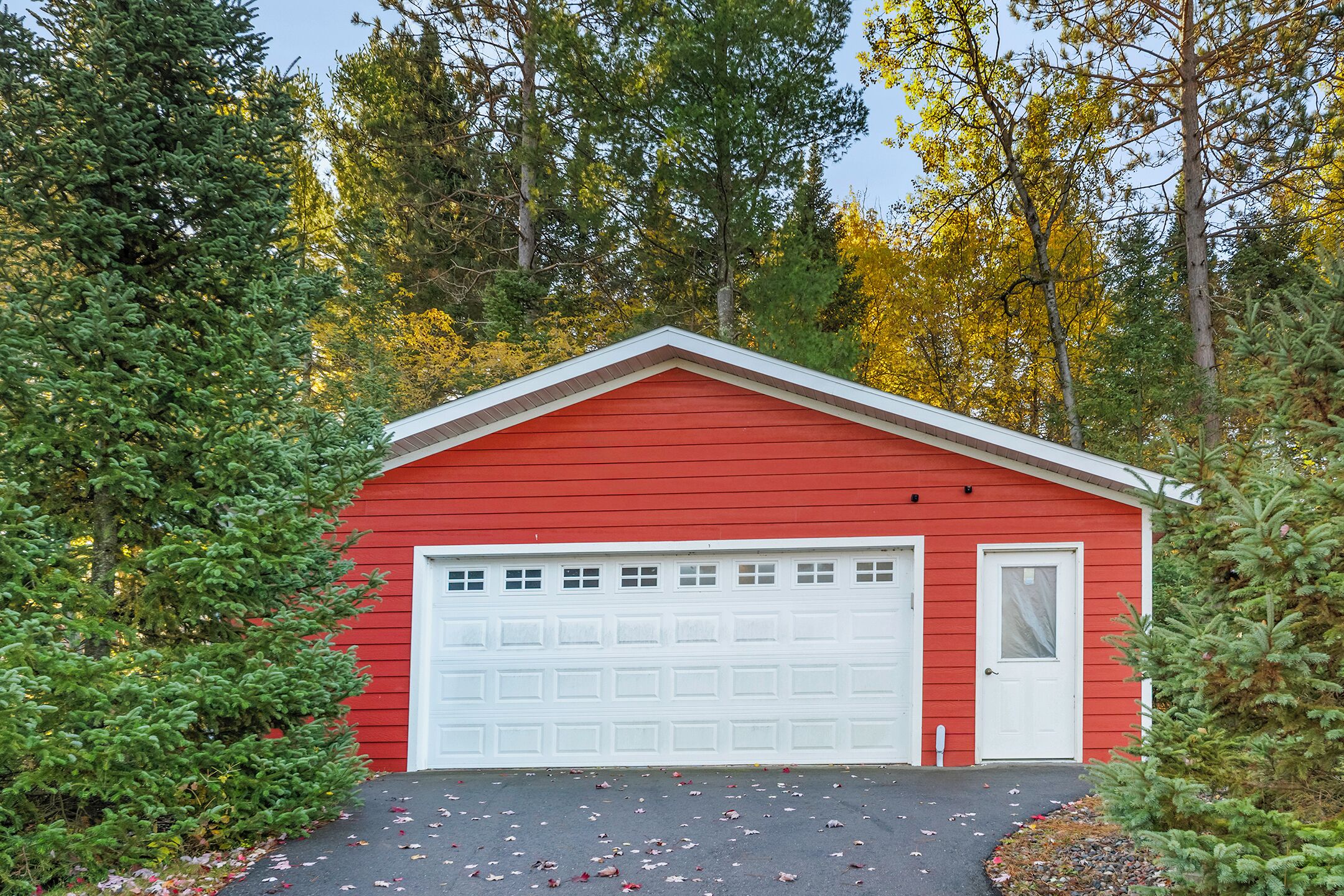 Un garage rouge avec une porte et un portail de garage blancs, entouré d’arbres et de feuilles mortes.