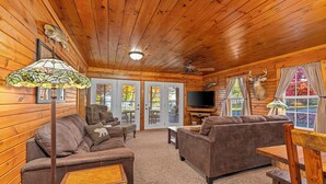 A cozy log cabin interior with a wooden ceiling, a large television, and a deer mounted on the wall.