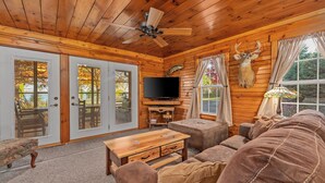 A log cabin interior with a wooden ceiling, a ceiling fan, a television, a sofa, and a deer head mounted on the wall.