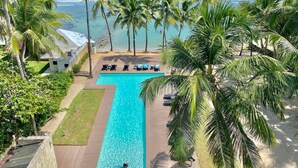 A resort pool with a view of the ocean, surrounded by palm trees and lounge chairs. - Beachfront 2BR Condo in Juan Dolio – Stylish, Cozy & Fully Equipped Retreat (Playa Juan Dolio)