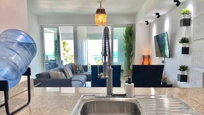 A modern kitchen with a stainless steel sink, a hanging light fixture, and a view of a living room with a sofa and a television. - Beachfront 2BR Condo in Juan Dolio – Stylish, Cozy & Fully Equipped Retreat (Playa Juan Dolio)