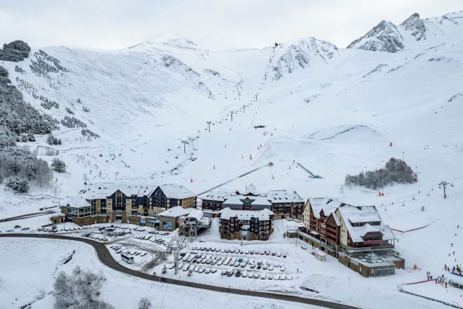 Un centro turístico en la montaña cubierto de nieve, con varios edificios, telesillas y un aparcamiento.
