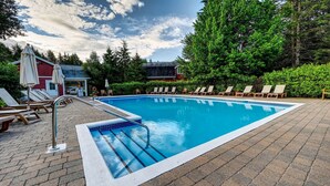 Una piscina rectangular con borde de azulejos, rodeada de sillas de descanso y sombrillas, con un fondo de árboles y un edificio con techo rojo.
