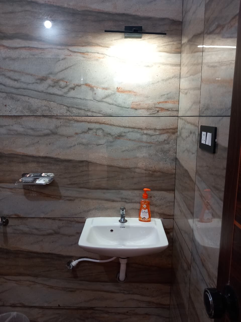 A bathroom with a marble wall, a sink with a soap dispenser, and a light fixture above the mirror.