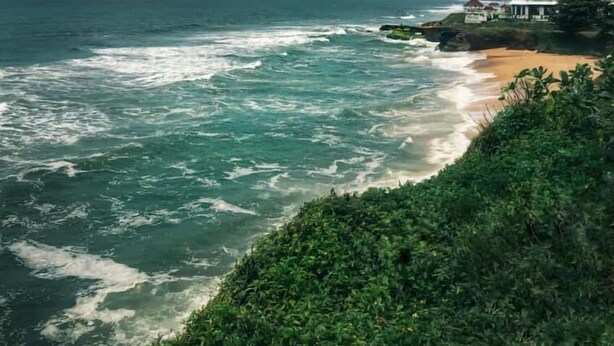 Vista costera con olas rompiendo sobre una playa arenosa, un edificio de techo rojo y abundante vegetación.