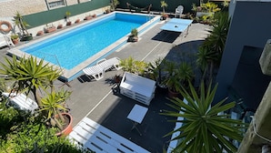 A swimming pool with lounge chairs and potted plants, surrounded by buildings and greenery. - Slide Surfcamp (Vagos)