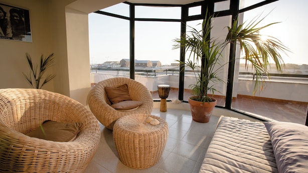 A spacious balcony with wicker furniture, a potted plant, and a view of the cityscape. - Slide Surfcamp (Vagos)