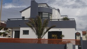 A modern two-story house with a curved balcony, a small garden, and a clear blue sky. - Slide Surfcamp (Vagos)