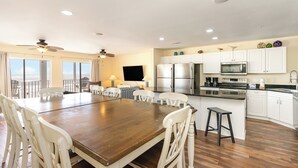 A spacious dining area with a large wooden table and white chairs, a kitchen with white cabinets and stainless steel appliances, and a balcony with a view. - Ambassador Villas 401 Oceanfront 7 Br 6 Ba (North Myrtle Beach)