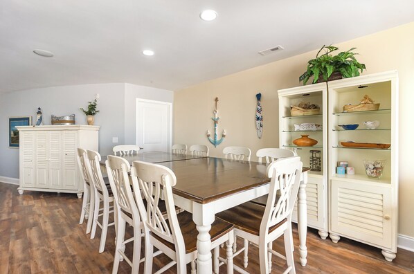 Un coin repas avec une table en bois et des chaises blanches, un meuble blanc avec des étagères en verre, et une porte visible au fond.