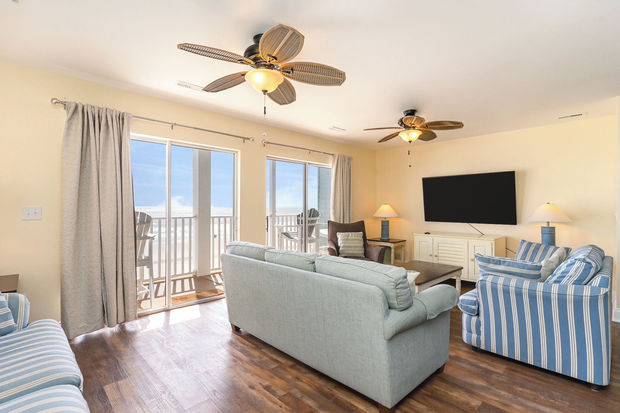 A spacious living room with a large sofa, striped armchairs, a flat-screen TV, ceiling fans, and a balcony with a view.