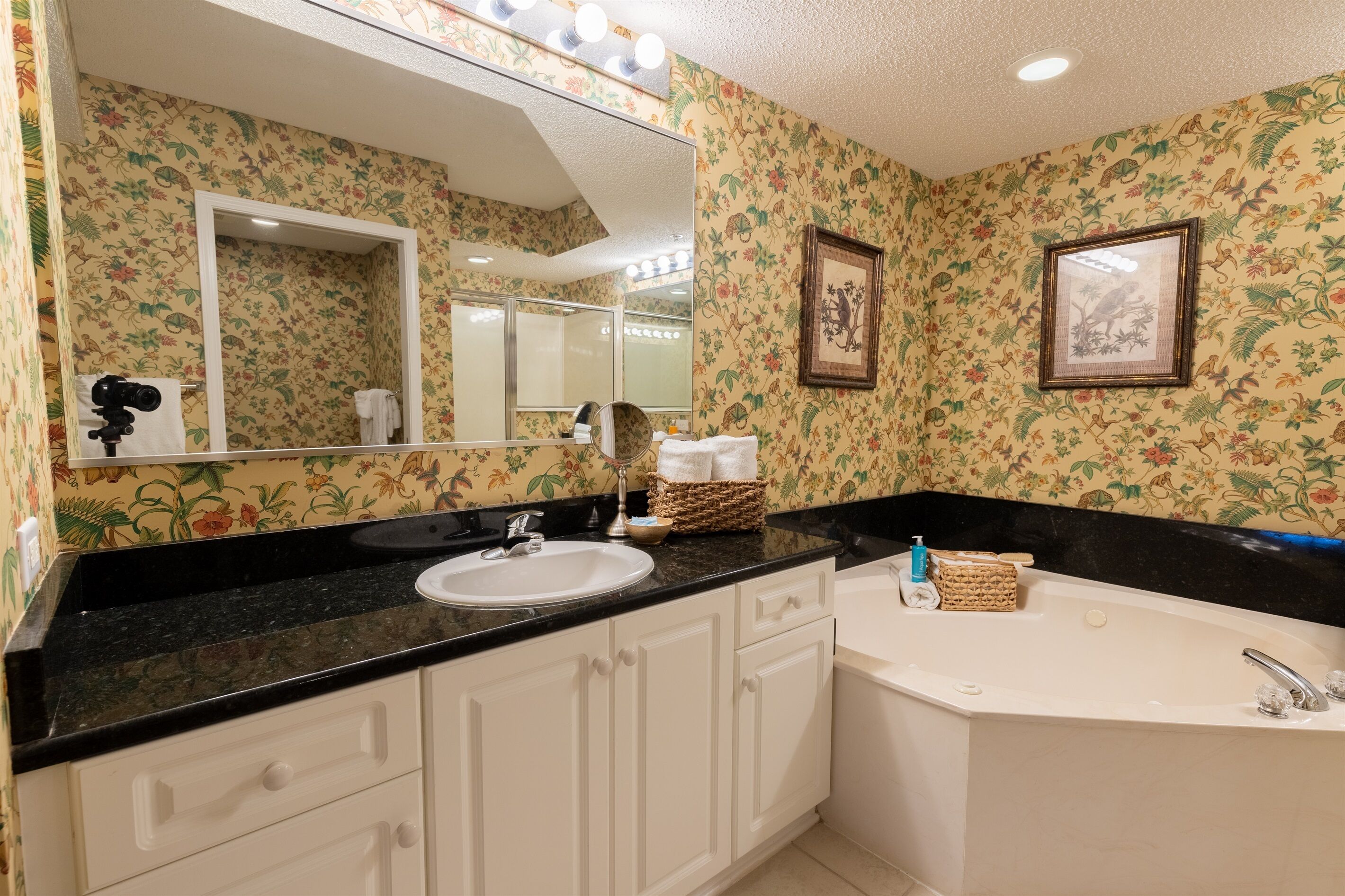 Un baño con papel tapiz floral, un espejo grande, un lavabo, una bañera y un tocador con encimera negra.