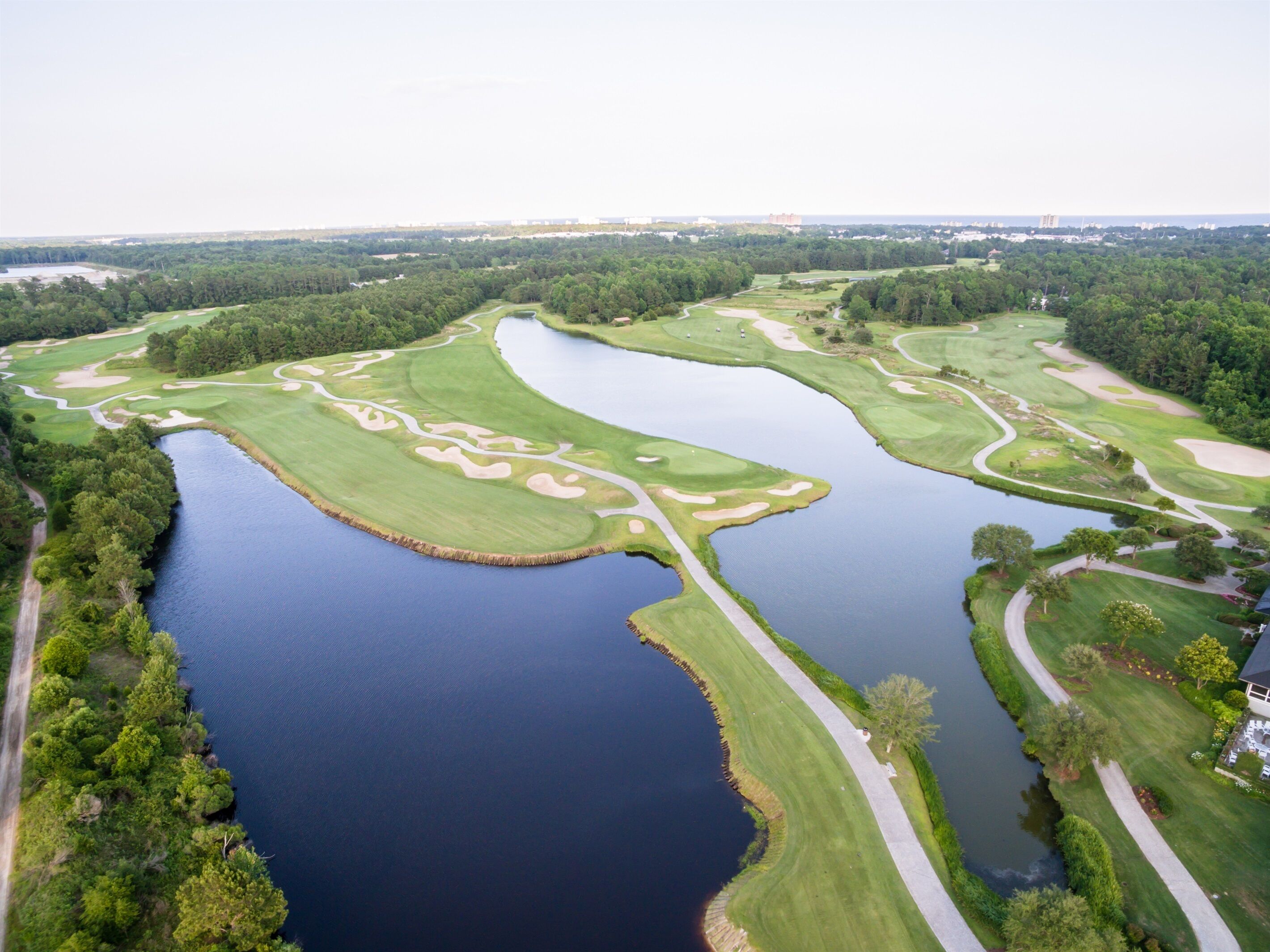 Golfkenttä, jossa on useita vesiesteitä ja hiekkabunkkereita, ja sen ympärillä on tiheää puustoa sekä avoin, kirkas taivas.