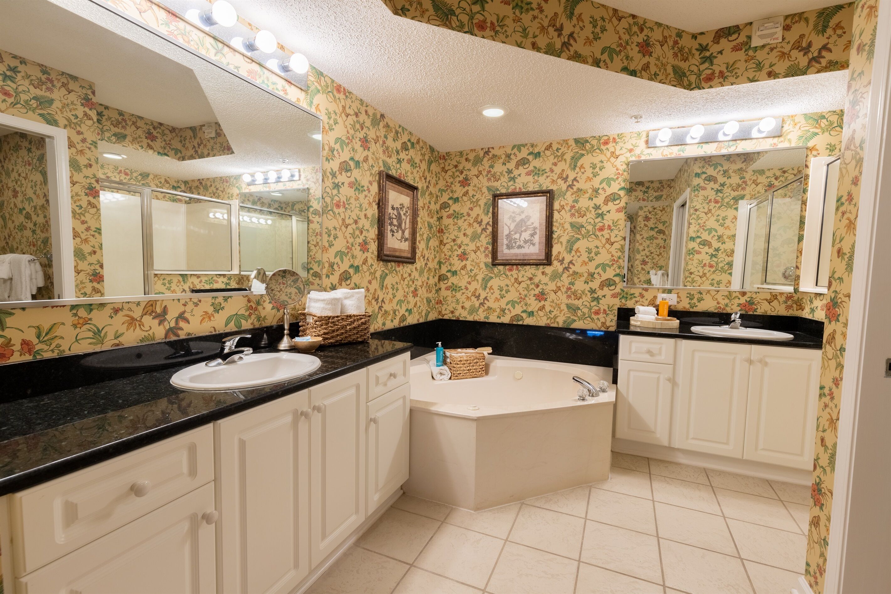 Un baño con papel tapiz floral, encimera negra, lavamanos blanco, bañera y un espejo grande.