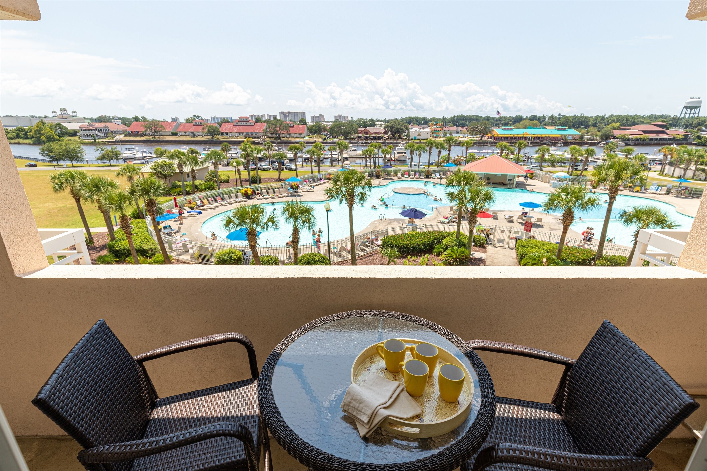 A balcony with wicker chairs and a table, overlooking a pool area with palm trees and lounge chairs.