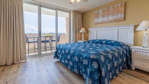 A bedroom with a large bed, a ceiling fan, a window with curtains, a painting on the wall, and a nightstand with a lamp. - Barefoot N Tower 404- Golf, Pool, Free Shuttle (North Myrtle Beach)