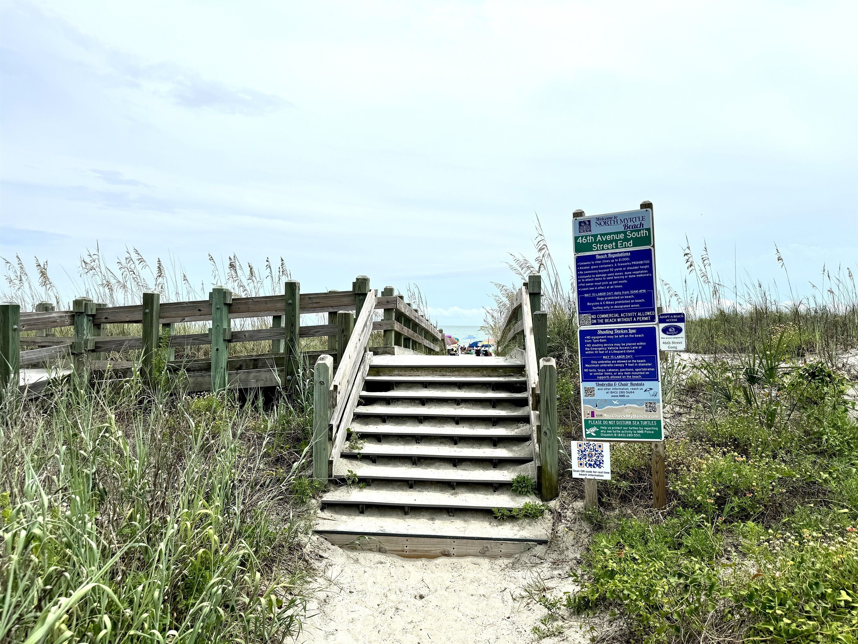 Un escalier en bois mène à une zone de plage où se trouve un panneau indiquant les règles à respecter sur la plage, ainsi qu'un code QR pour obtenir de plus amples informations.