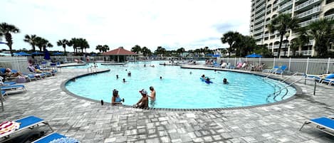 A swimming pool with lounge chairs and umbrellas, surrounded by palm trees and a hotel building.