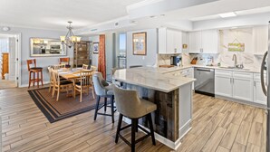 A modern kitchen with a central island, white cabinetry, and a dining area with a wooden table and chairs. - Brighton 1410 3 Br Oceanfront in Kingston w/Pool (Myrtle Beach)
