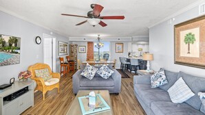 A spacious living room with a gray sofa, a wicker chair, a coffee table, and a ceiling fan. - Brighton 1410 3 Br Oceanfront in Kingston w/Pool (Myrtle Beach)