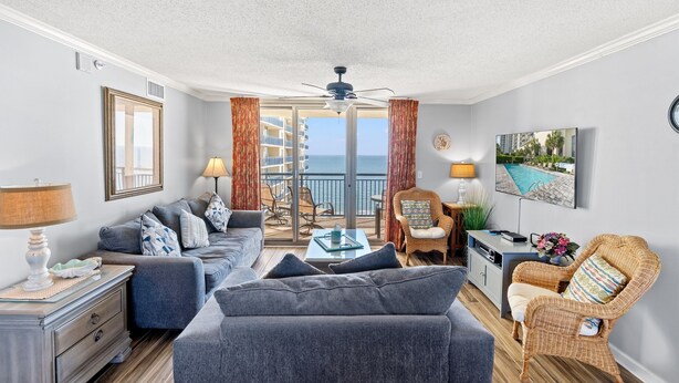 A modern living room with a grey sofa, a wicker chair, a small table, and a balcony with a view of the sea. - Brighton 1410 3 Br Oceanfront in Kingston w/Pool (Myrtle Beach)