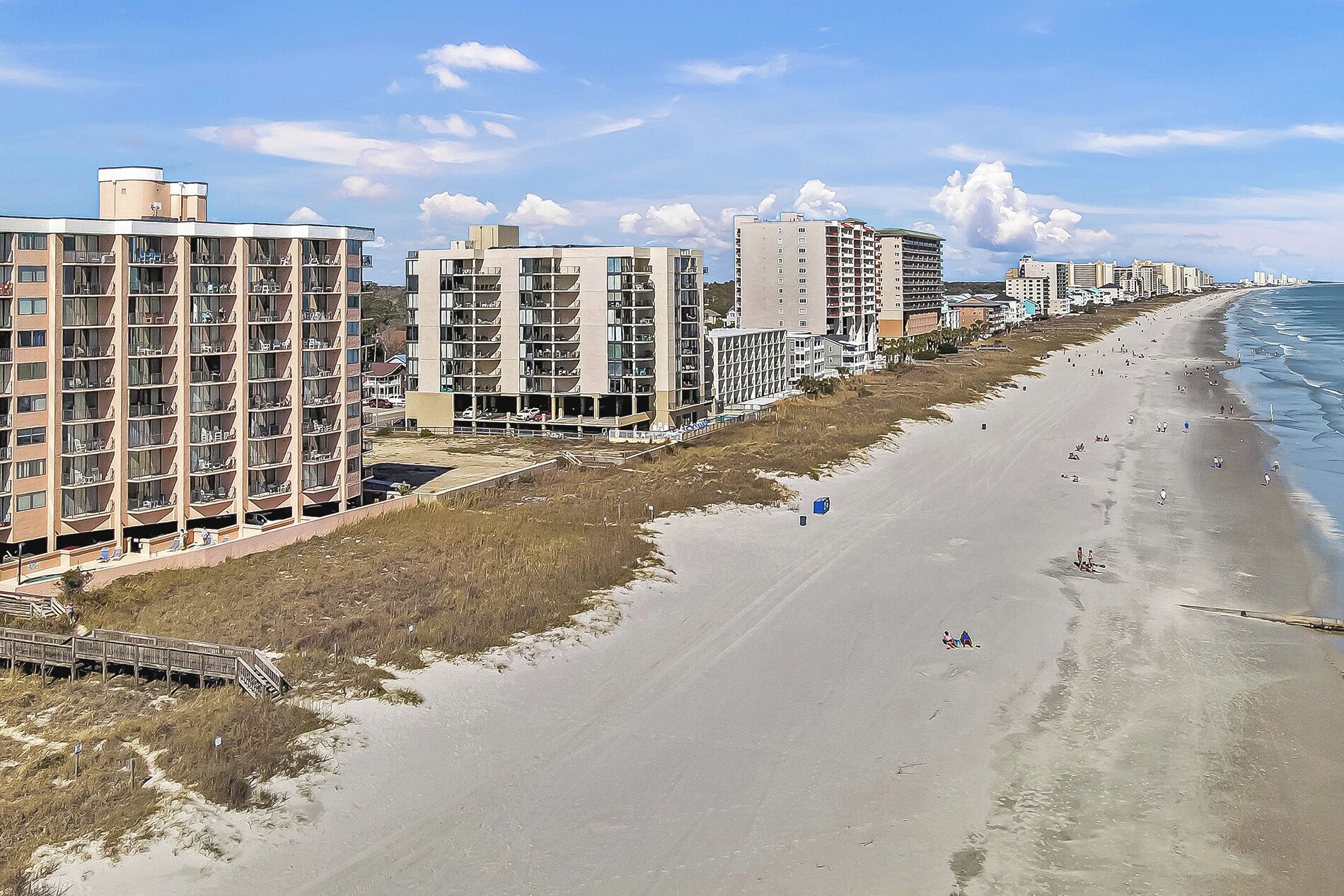 A beachfront hotel with multiple floors, a boardwalk leading to the beach, and a sandy shoreline with a few people.
