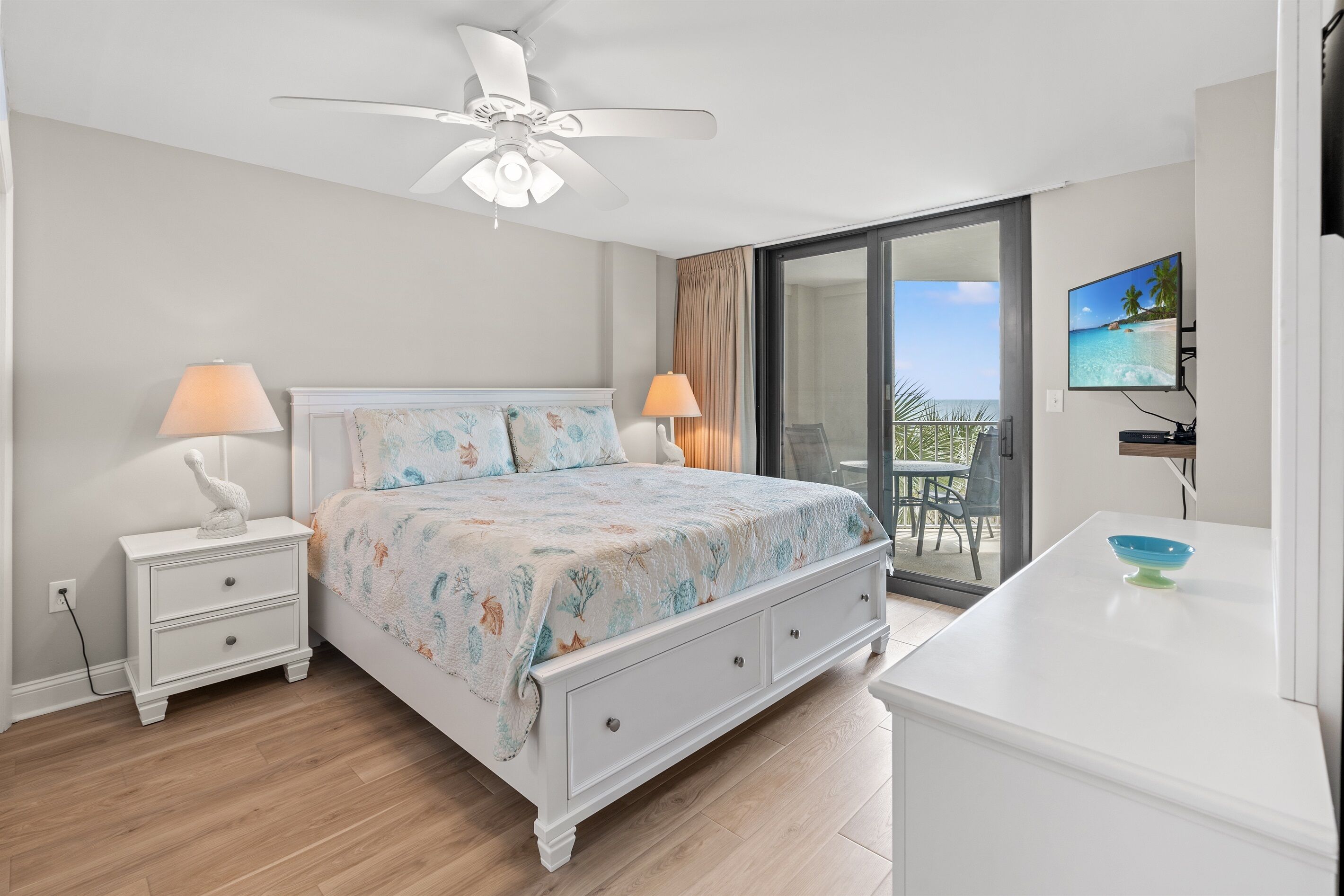 Ein Schlafzimmer mit einem Bett, Nachttischen, einem Schreibtisch, einem Deckenventilator, einem Fernseher und einer Schiebetür zum Balkon.
