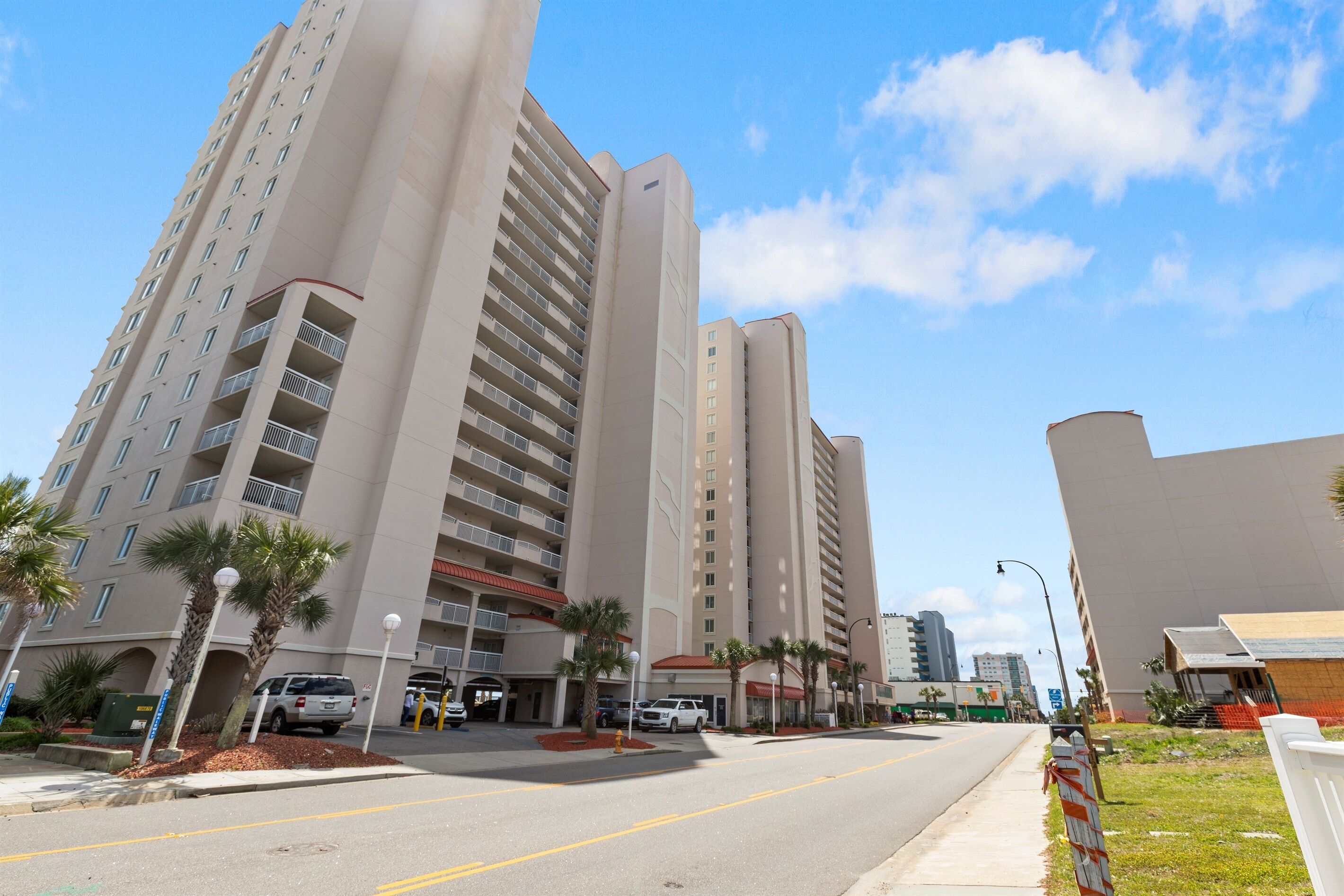 A multi-story hotel with balconies, a parking area, and palm trees.
