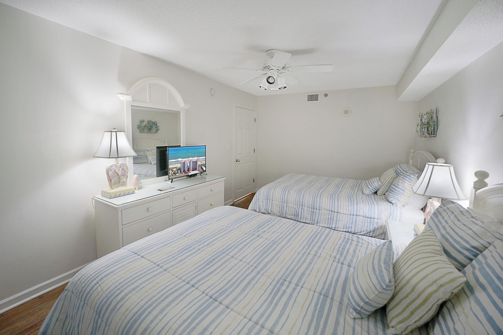 Une chambre avec deux lits, un ventilateur de plafond, un téléviseur posé sur une commode et un miroir.