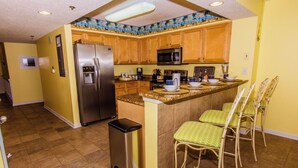 A kitchen with wooden cabinets, a stainless steel refrigerator, a microwave, and a granite countertop. There are two bar stools with green cushions.