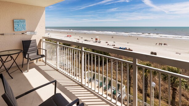 A balcony with a view of the beach, a table, two chairs, and a sign with beach rules.