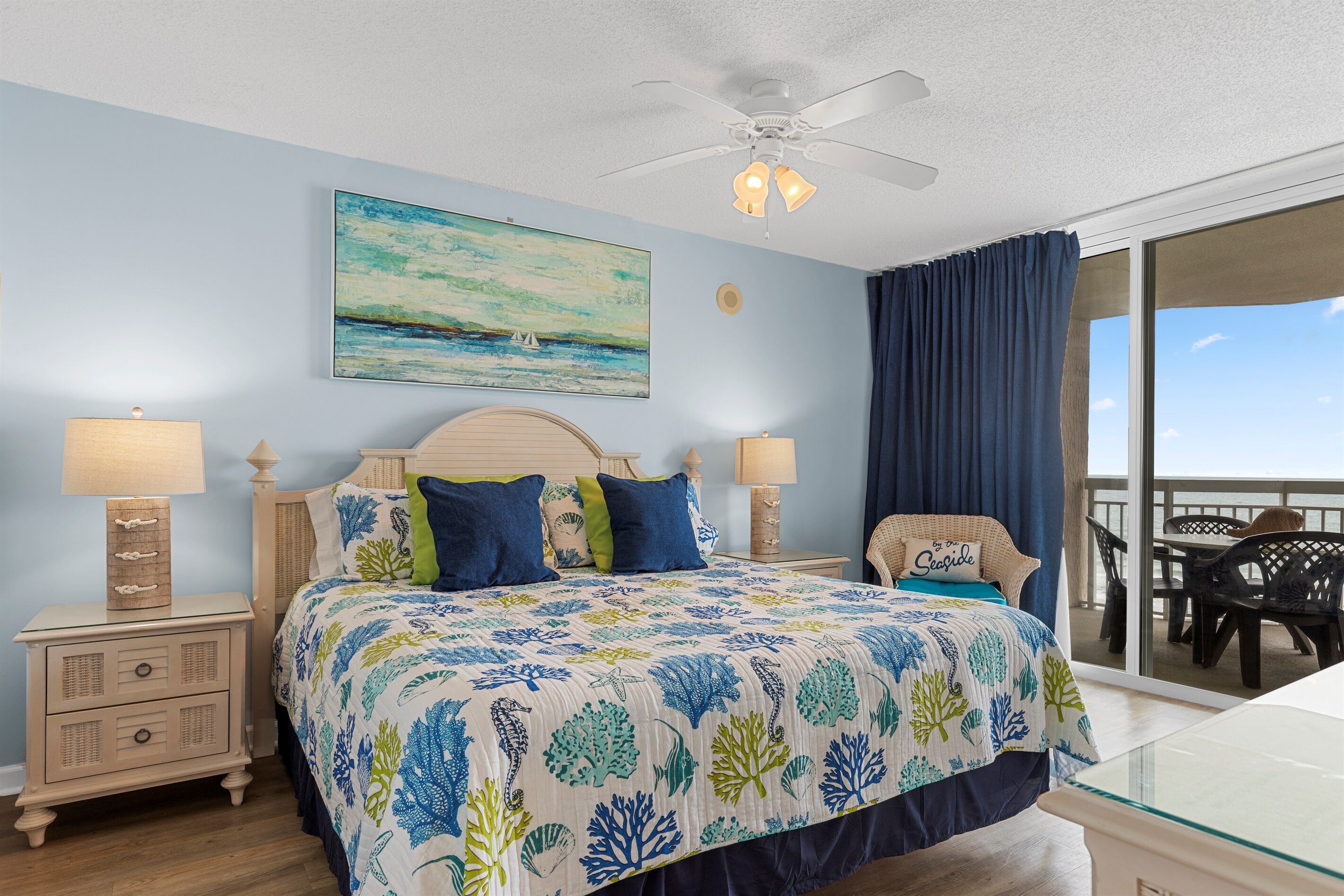 A bedroom with a bed, two bedside tables, a ceiling fan, a painting, and a view of the outdoors.