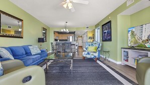 A living room with a blue sofa, a coffee table, and a television on a stand. There is a ceiling fan and a kitchen in the background. - Crescent Shores 807 Oceanfront, Pools, Lazy River (North Myrtle Beach)