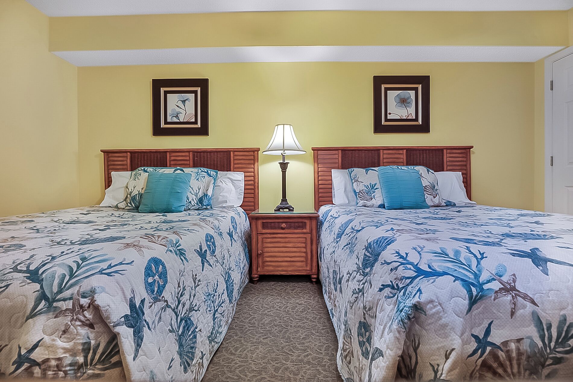 A hotel room with two beds, a nightstand, a lamp, and two framed pictures on the wall.