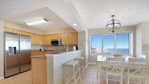 A modern kitchen with a central island, stainless steel appliances, wooden cabinets, and a dining area with white chairs. - Crescent Shores 1601 Oceanfront, Pools,Lazy River (North Myrtle Beach)