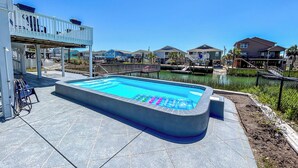 Una piscina rectangular con jacuzzi, rodeada por una terraza con sillas y una pérgola.
