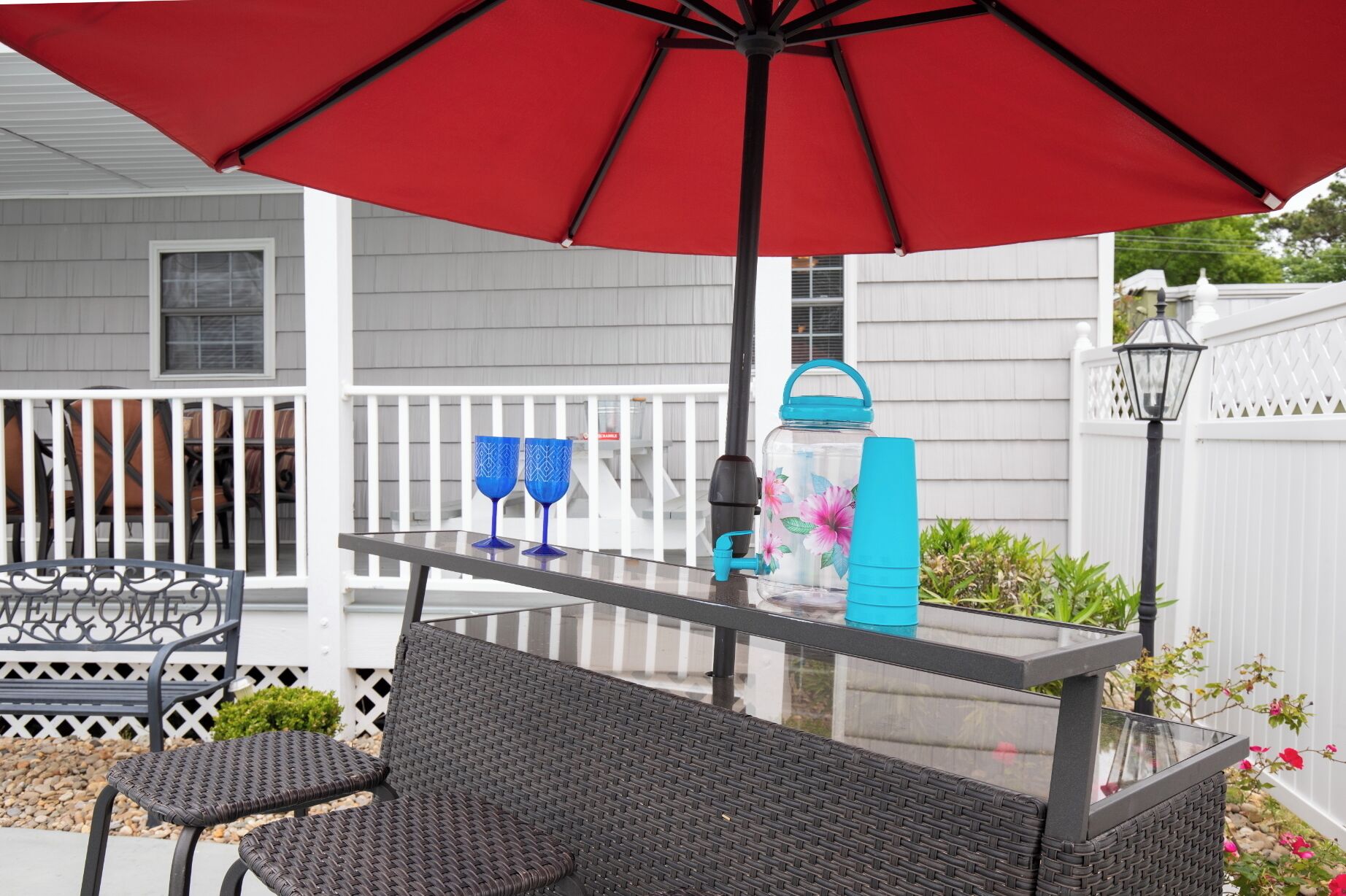 Une terrasse extérieure avec une table en verre, deux fauteuils en osier, un parasol rouge et une bouteille d’eau.