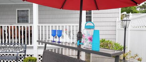 Une terrasse extérieure avec une table en verre, deux fauteuils en osier, un parasol rouge et une bouteille d’eau.