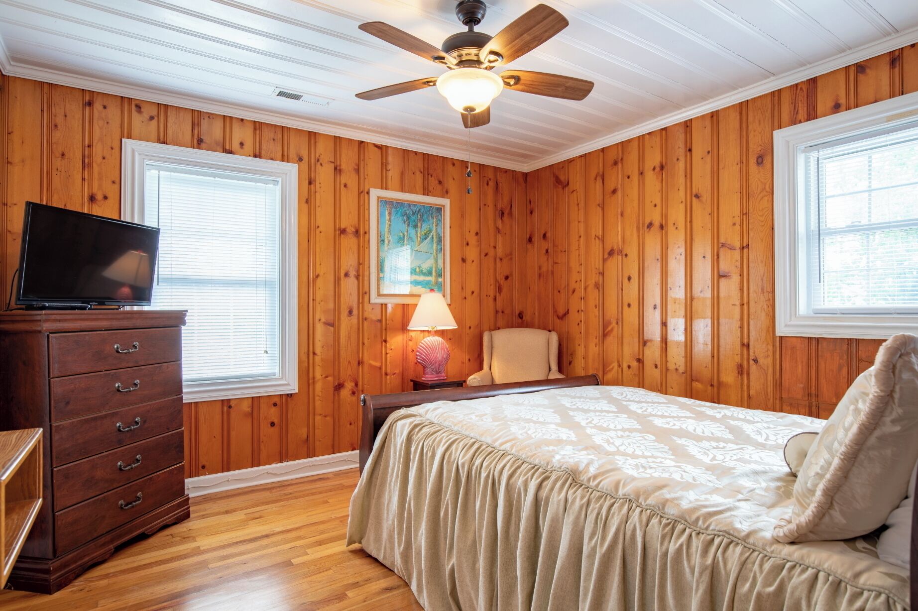 A bedroom with wooden paneling, a ceiling fan, a bed with a beige bedspread, a nightstand with a lamp, and a TV on a dresser.