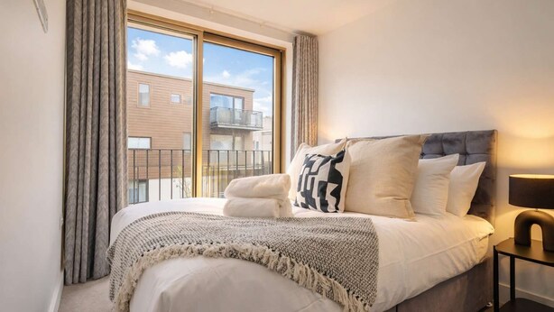 Un dormitorio con cama, mesita de noche, lámpara y vista a un edificio desde la ventana.